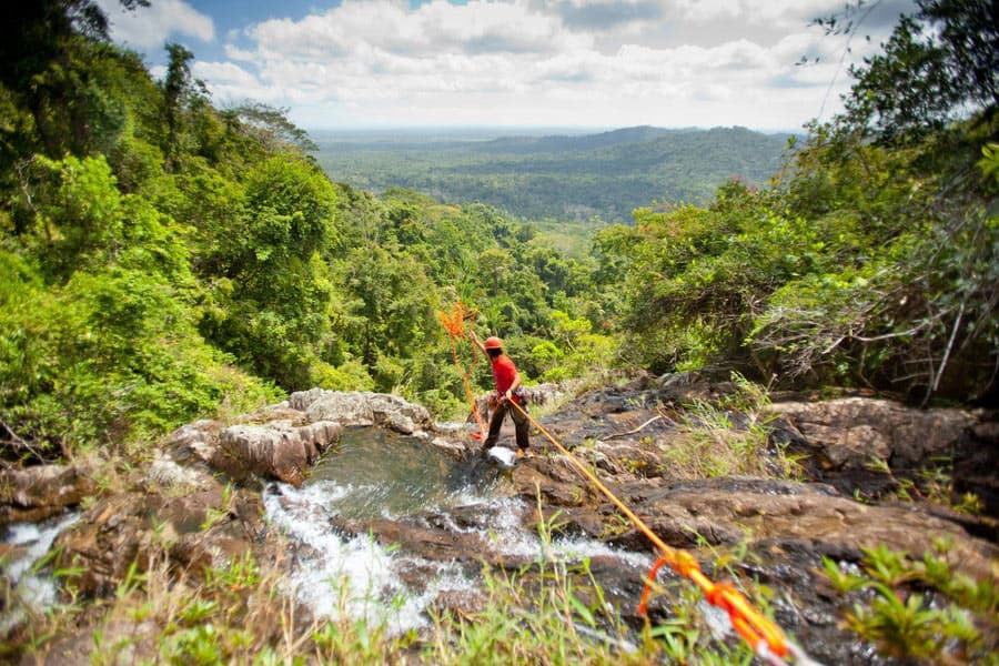Man on top of waterfalls with the jungle in the background throwing rappelling rope over the side of the cliff.