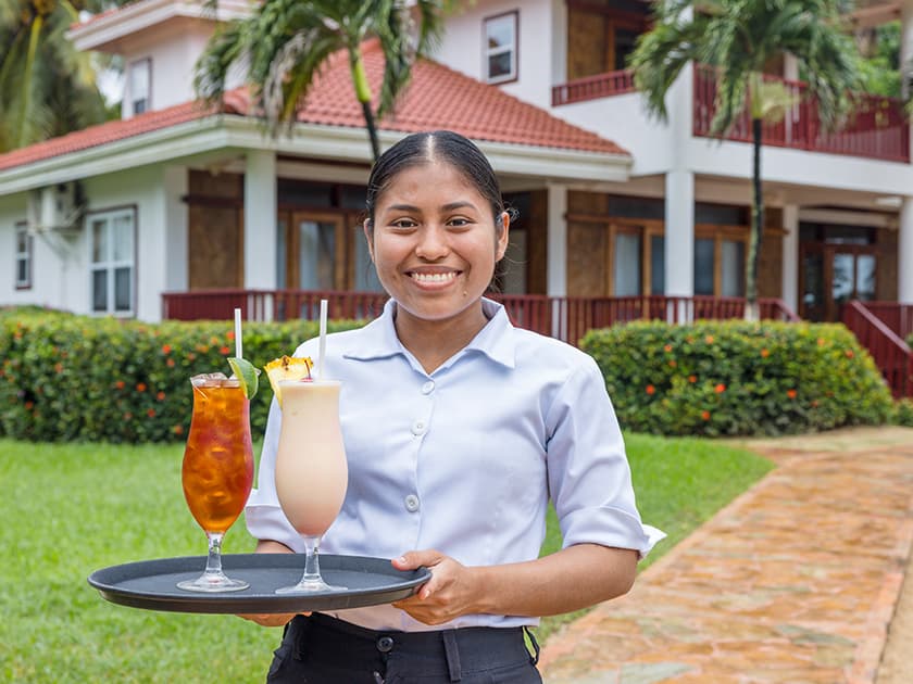 A smiling server holds a tray with two colorful cocktails in front of a tropical building. A smiling server holds a tray with two colorful cocktails in front of a tropical building.