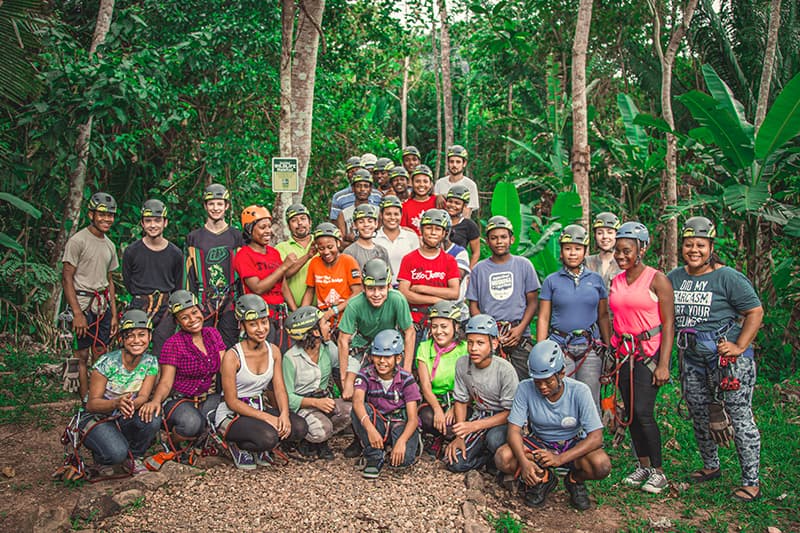 A diverse group of young people and adults wearing helmets and harnesses poses together in a lush, green forest.