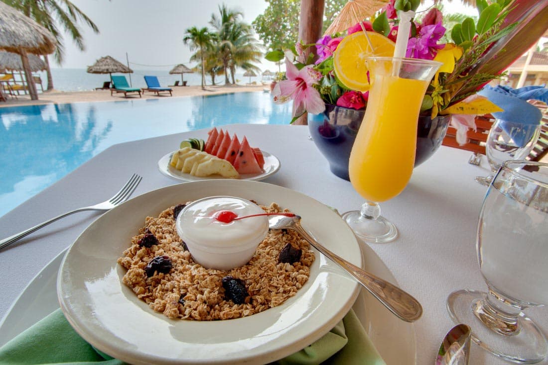 A delicious breakfast spread on a table overlooking a pool at Belizean Dreams Resort. The meal includes a bowl of granola with yogurt and a cherry, a plate of sliced fruits, a glass of orange juice, and a glass of water. The pool is surrounded by lounge chairs and palm trees.