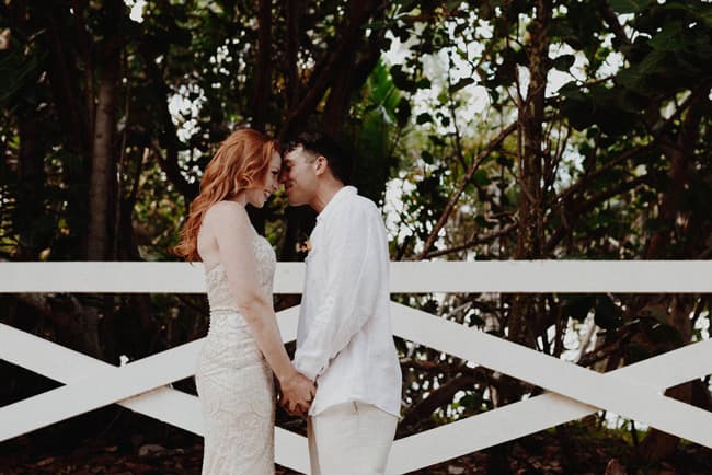 A couple in wedding attire leans in close, holding hands against a white fence with lush greenery in the background.