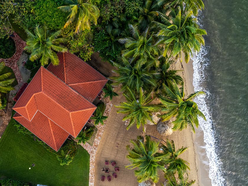 Aerial view of a coastal house surrounded by palm trees and sandy beach.