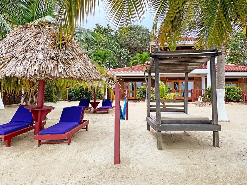 A sandy beach area featuring purple lounge chairs, a thatched umbrella, and a shaded structure surrounded by tropical greenery. A sandy beach area featuring purple lounge chairs, a thatched umbrella, and a shaded structure surrounded by tropical greenery.