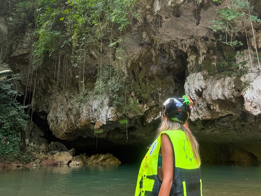 A person wearing a bright yellow life vest and helmet gazes up at a rocky, moss-covered cave entrance surrounded by lush greenery. A person wearing a bright yellow life vest and helmet gazes up at a rocky, moss-covered cave entrance surrounded by lush greenery.