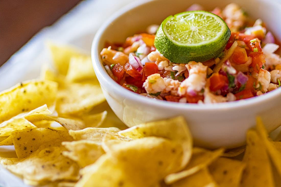 A white bowl filled with ceviche, a colorful seafood dish made with shrimp, tomatoes, onions, and other ingredients. A lime wedge sits on top of the ceviche, and a pile of tortilla chips is scattered around the bowl.