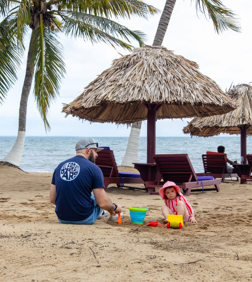 A man sits on the beach with a child playing in the sand near thatched umbrellas.