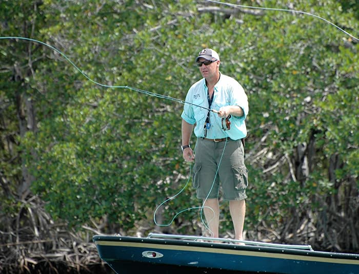 A man in a ball cap stands in a boat casting a fly fishing line into the river.