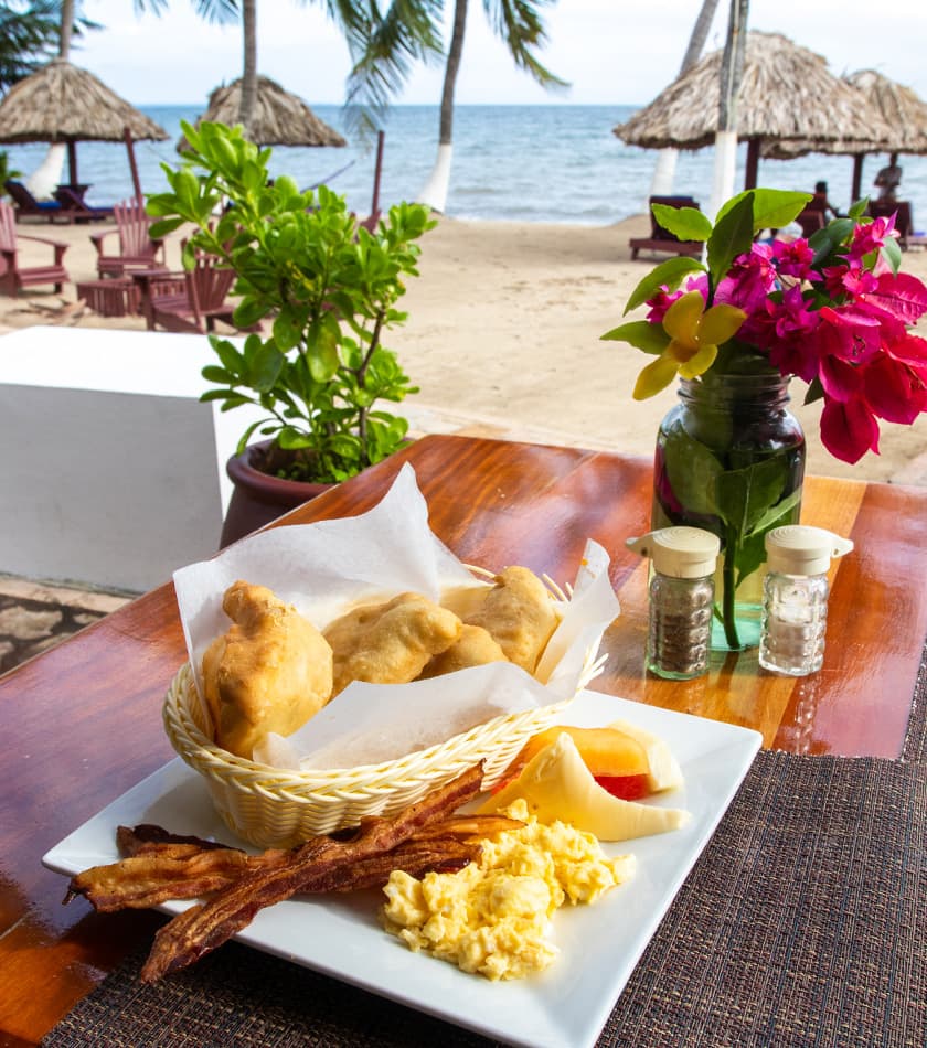 A plate of breakfast featuring fried pastries, scrambled eggs, and fruit, with a beach view in the background.