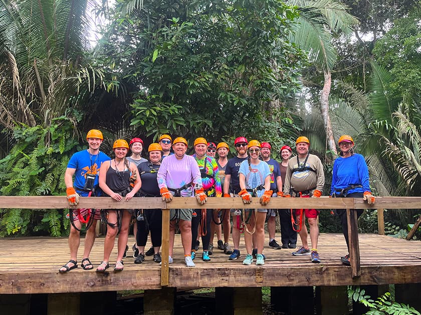 A group of people wearing helmets and safety gear stands on a wooden platform in a lush, tropical forest.