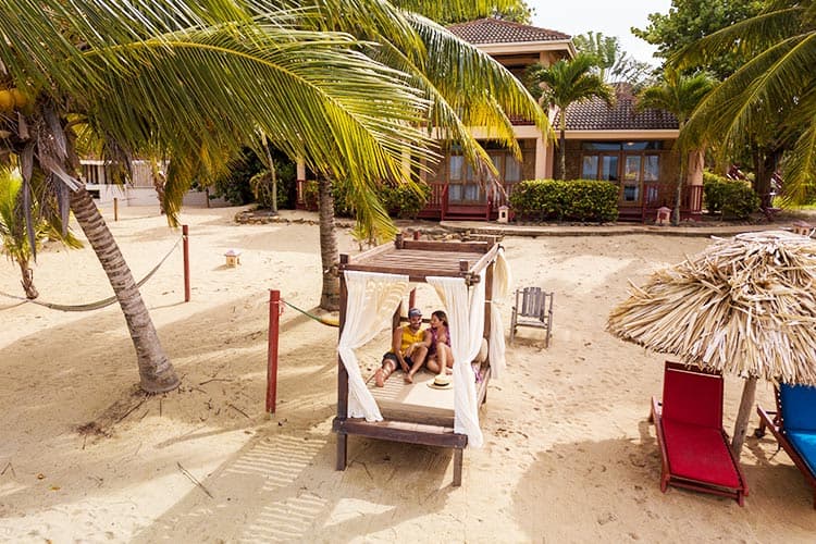 A family enjoys a cozy moment on a shaded beach cabana surrounded by palm trees.