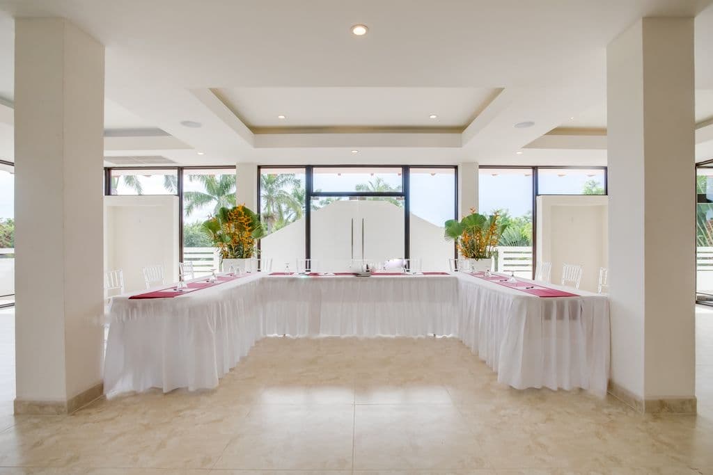 A spacious and modern meeting room with a U-shaped table arrangement. The room has a white tiled floor, white walls, and a high ceiling with exposed beams. Large windows offer views of a green outdoor space. The table is covered in white tablecloths and has red runners. Clear plastic chairs are arranged around the table.