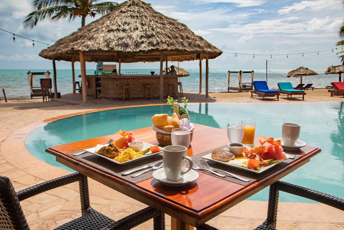 A picturesque beachfront scene at Belizean Dreams Resort. A thatched-roof bar overlooks a swimming pool and the ocean. Lounge chairs are arranged around the pool, and a wooden table with chairs is set up for outdoor dining.
