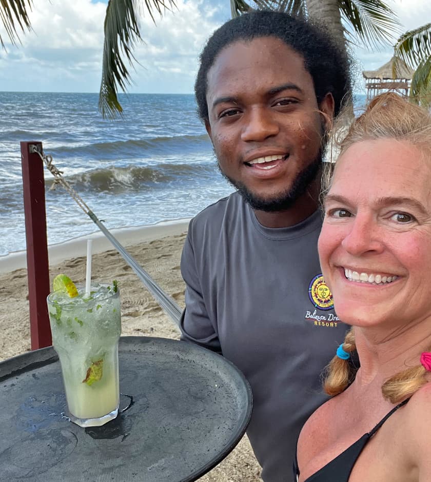 A smiling woman poses with a drink next to a server on a beach.