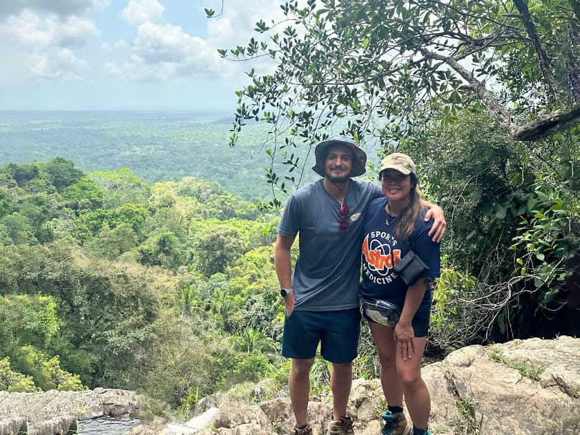 A man and a woman pose together on a rocky outcrop with a lush green landscape in the background.