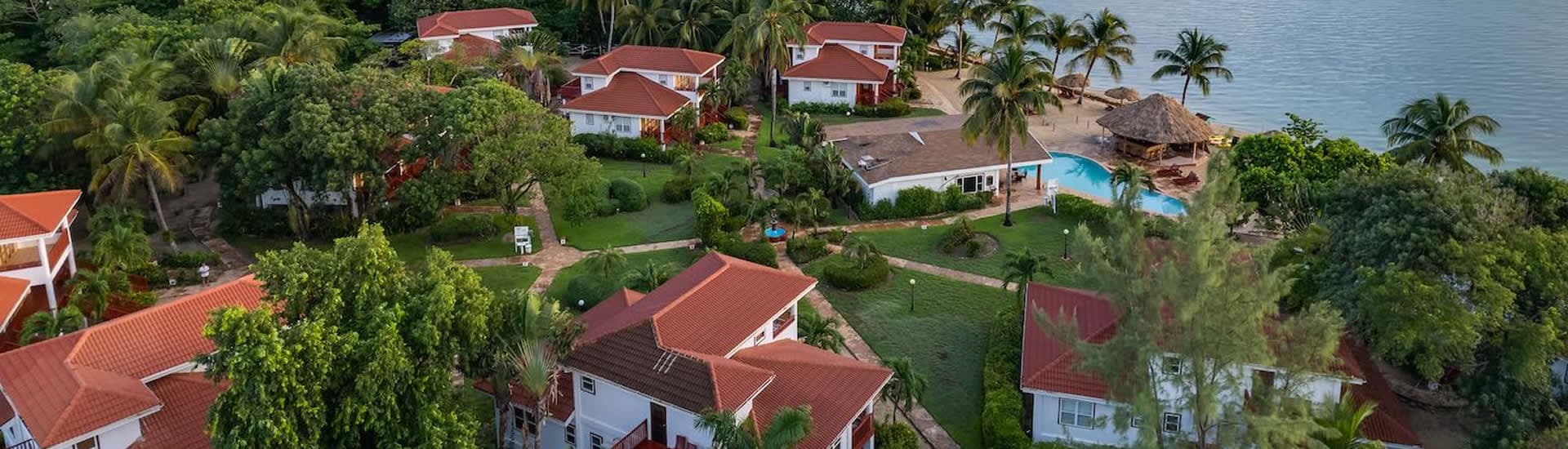 Ariel view of property, with red roof buildings, palm trees, green grass, pool and ocean in the background.