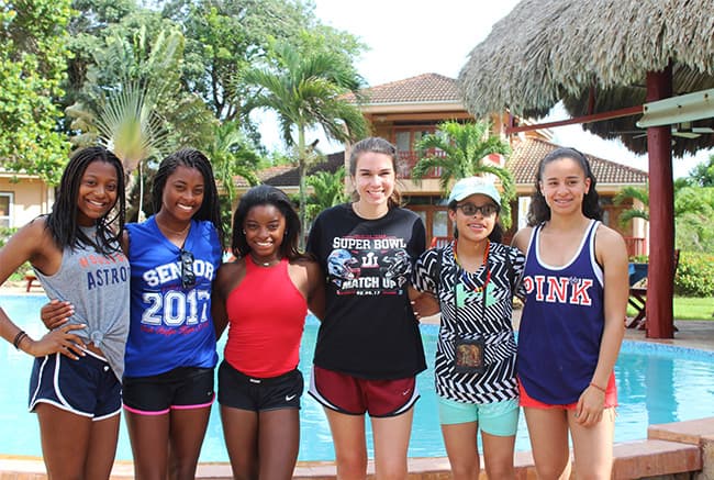 A group of six young women stands together by a pool, smiling and posing for the camera.