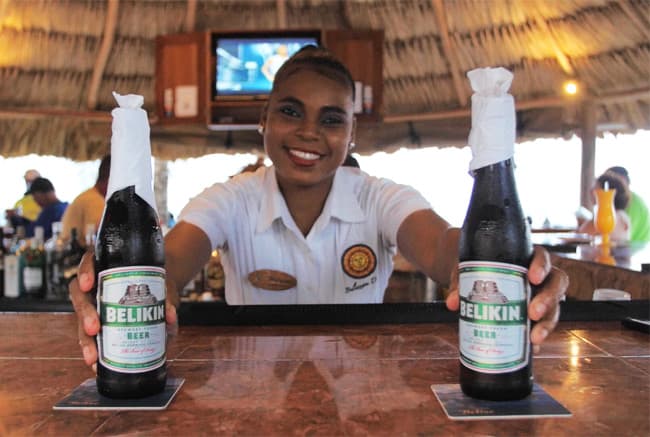 A smiling bartender presents two bottles of Belikin beer at a tropical bar.
