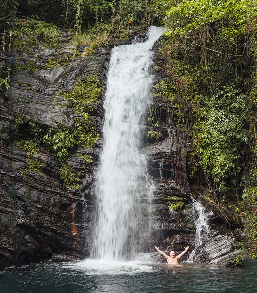 A person stands in a pool beneath a lush waterfall, with arms raised in celebration.