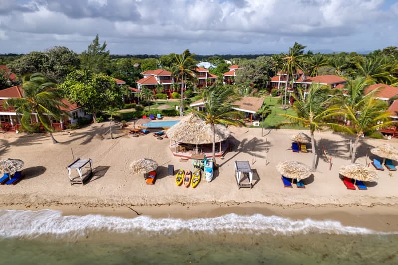 Aerial view of a sandy beach with thatched umbrellas, colorful lounge chairs, and resort buildings surrounded by palm trees.