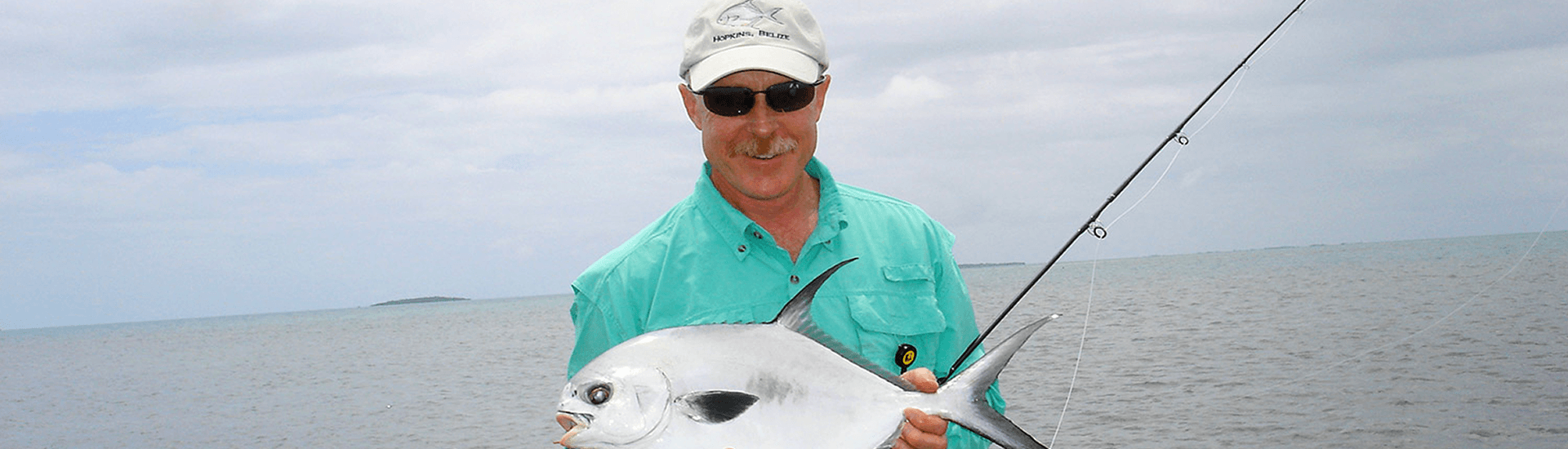 A man in a teal fishing shirt holds up his catch - a flat silvery fish.