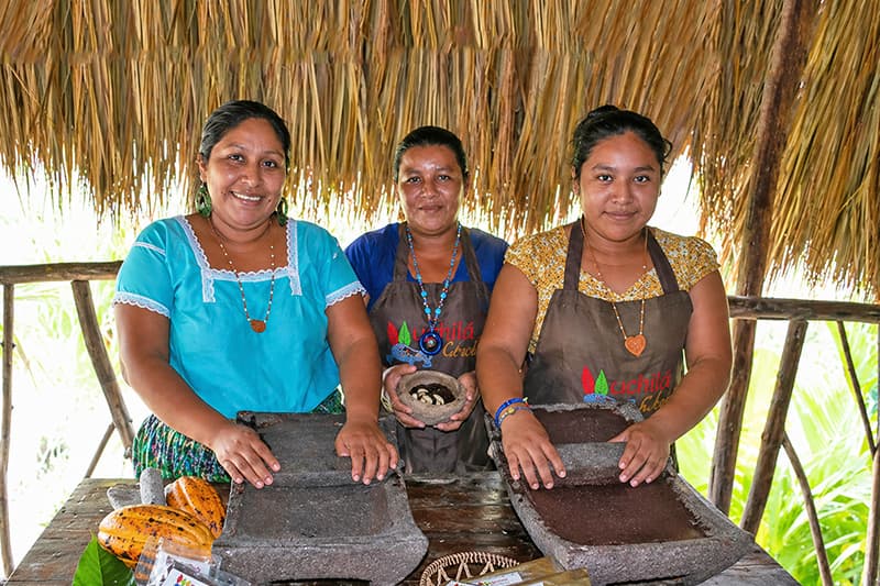 Three women in colorful traditional attire stand behind counters, preparing food in a thatched-roof setting. Three women in colorful traditional attire stand behind counters, preparing food in a thatched-roof setting.