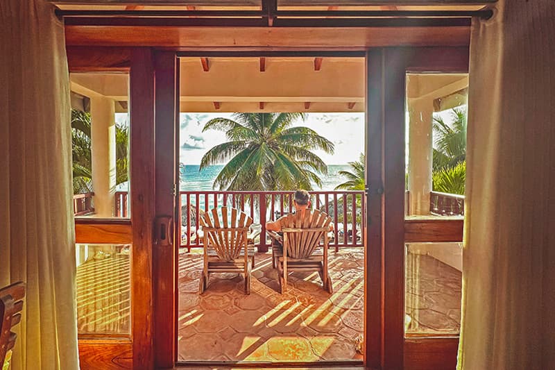 A person relaxes on a balcony overlooking the ocean, surrounded by palm trees.