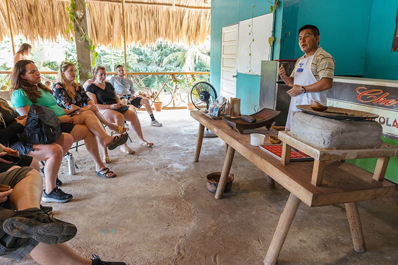 A chef demonstrates cooking techniques to an engaged audience seated around a wooden table in a tropical setting.