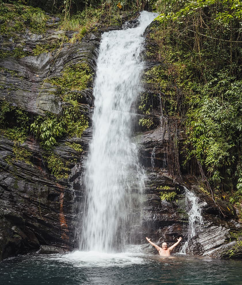 A person joyfully poses in a natural pool beneath a cascading waterfall surrounded by lush greenery.