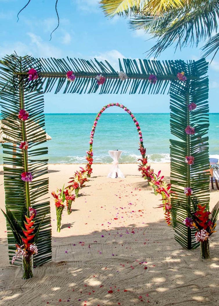 A tropical beach wedding setup with a floral archway made of palm leaves and colorful flowers. There is a white aisle runner leading to a small ceremony table, and the ocean and blue sky are visible in the background.