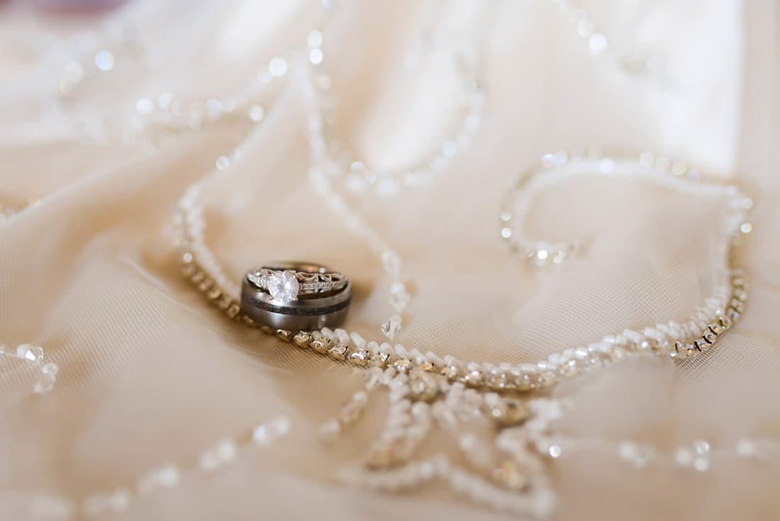 Two wedding rings resting on a beaded lace fabric. The engagement ring has a diamond, and the wedding band is made of metal.