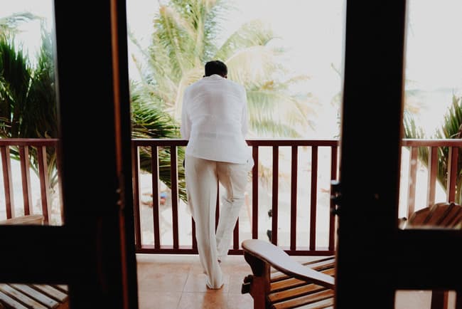 A person in white attire leans against a balcony railing, overlooking a palm-fringed landscape.