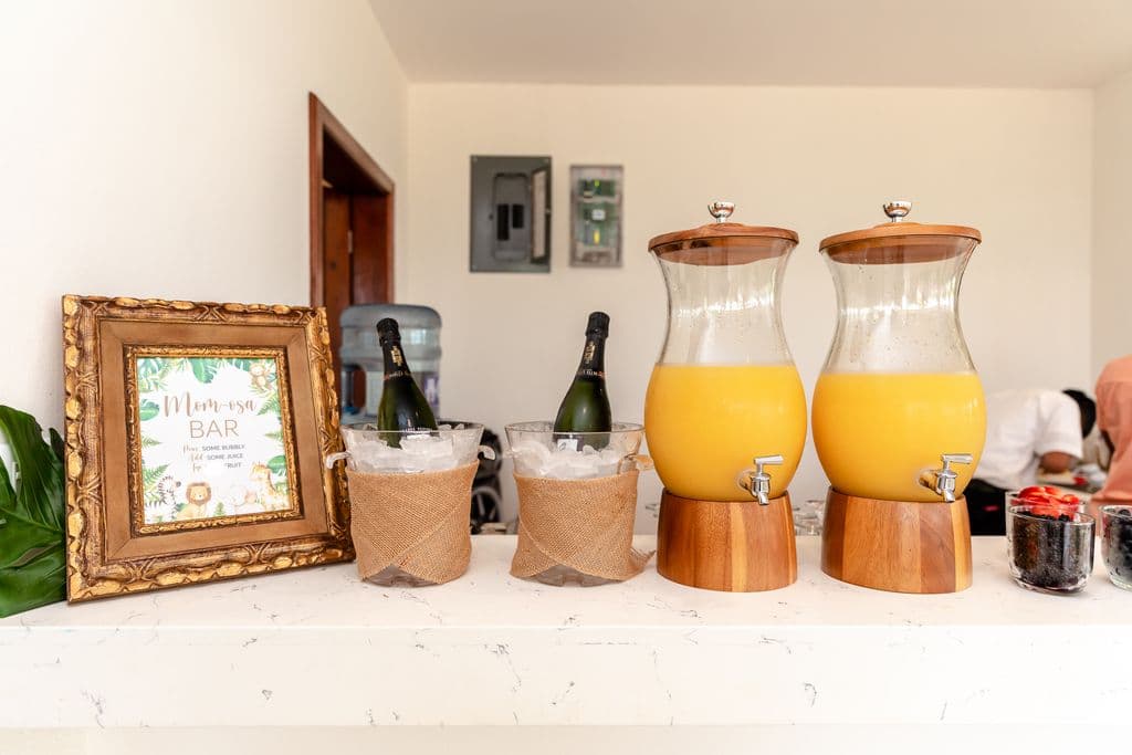 A "Mimosa Bar" sign is displayed in a gold frame above a bar counter. Two glass dispensers filled with orange juice and two bottles of champagne in ice buckets are on the counter.