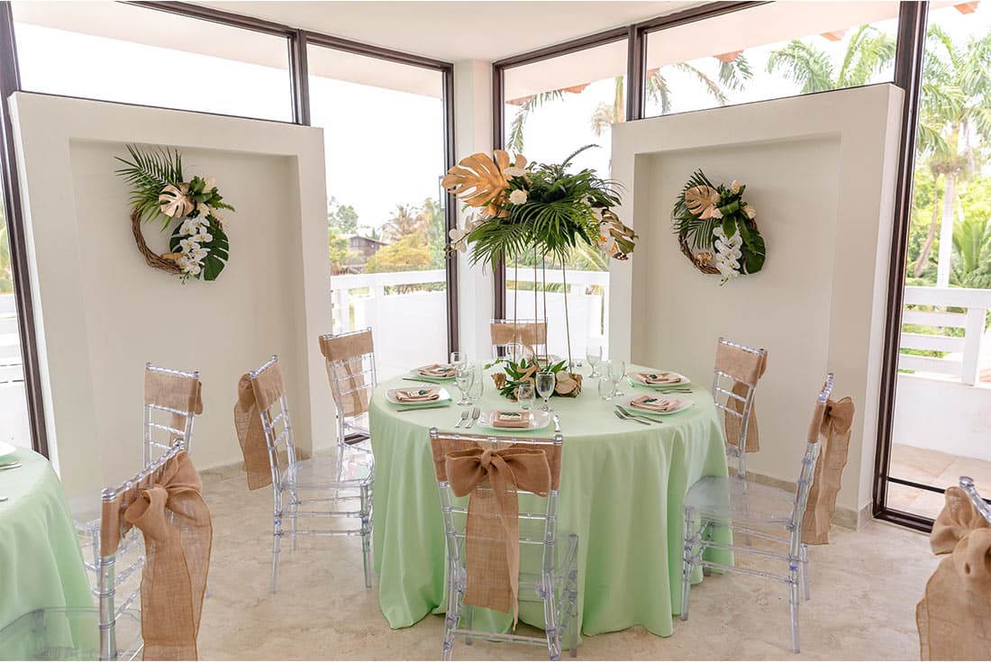 A beautifully decorated indoor wedding reception space with a round table covered in a mint green tablecloth and adorned with floral centerpieces. Clear ghost chairs with burlap sashes surround the table.