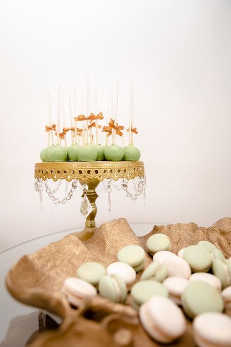 A gold cake stand holding a dozen green cake pops with orange bows. A gold bowl filled with green and white macarons sits in the foreground.