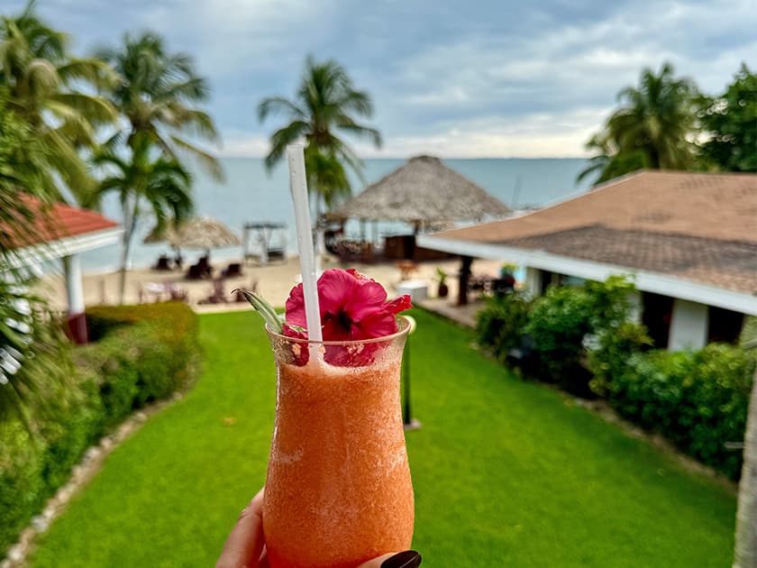 A tropical drink topped with a flower is held in front of a beachside resort. A tropical drink topped with a flower is held in front of a beachside resort.
