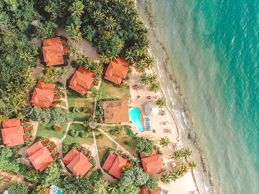 Aerial view of a coastal resort with red-roofed cabins, a swimming pool, and palm trees along the beach.
