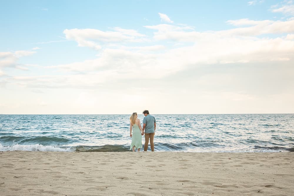 A woman in a green dress and a man in a blue shirt hold hands on a breezy day by the ocean's edge.