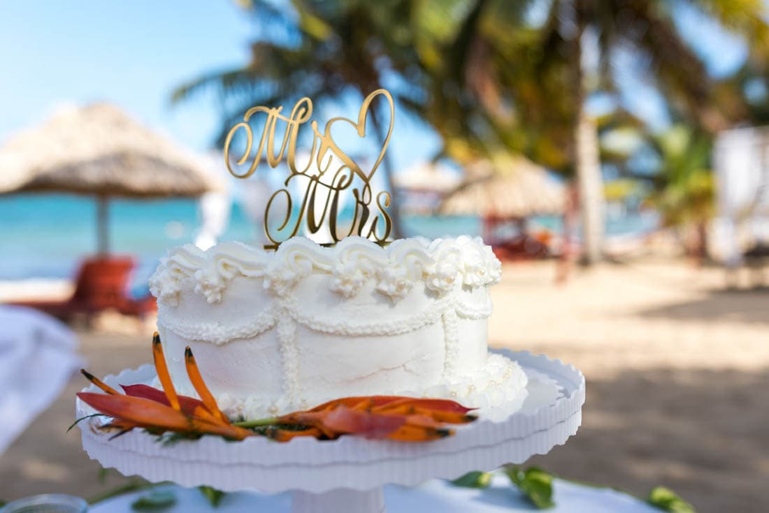 A white wedding cake decorated with buttercream frosting and a gold "Mrs. & Mrs." topper. The cake is placed on a white cake stand with orange flowers in front. The background shows a beach with blue water and palm trees.