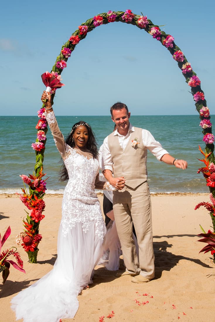 Ecstatic young bride and groom pose on the beach under a wedding arch decorated with pink, green and orange tropical flowers.