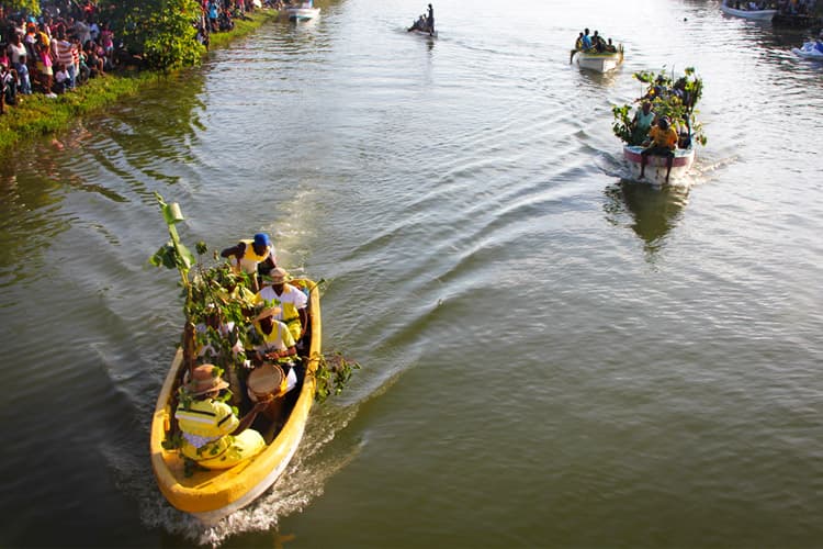 A group of people in decorated boats navigate a river during a vibrant festival, surrounded by spectators on the shore.