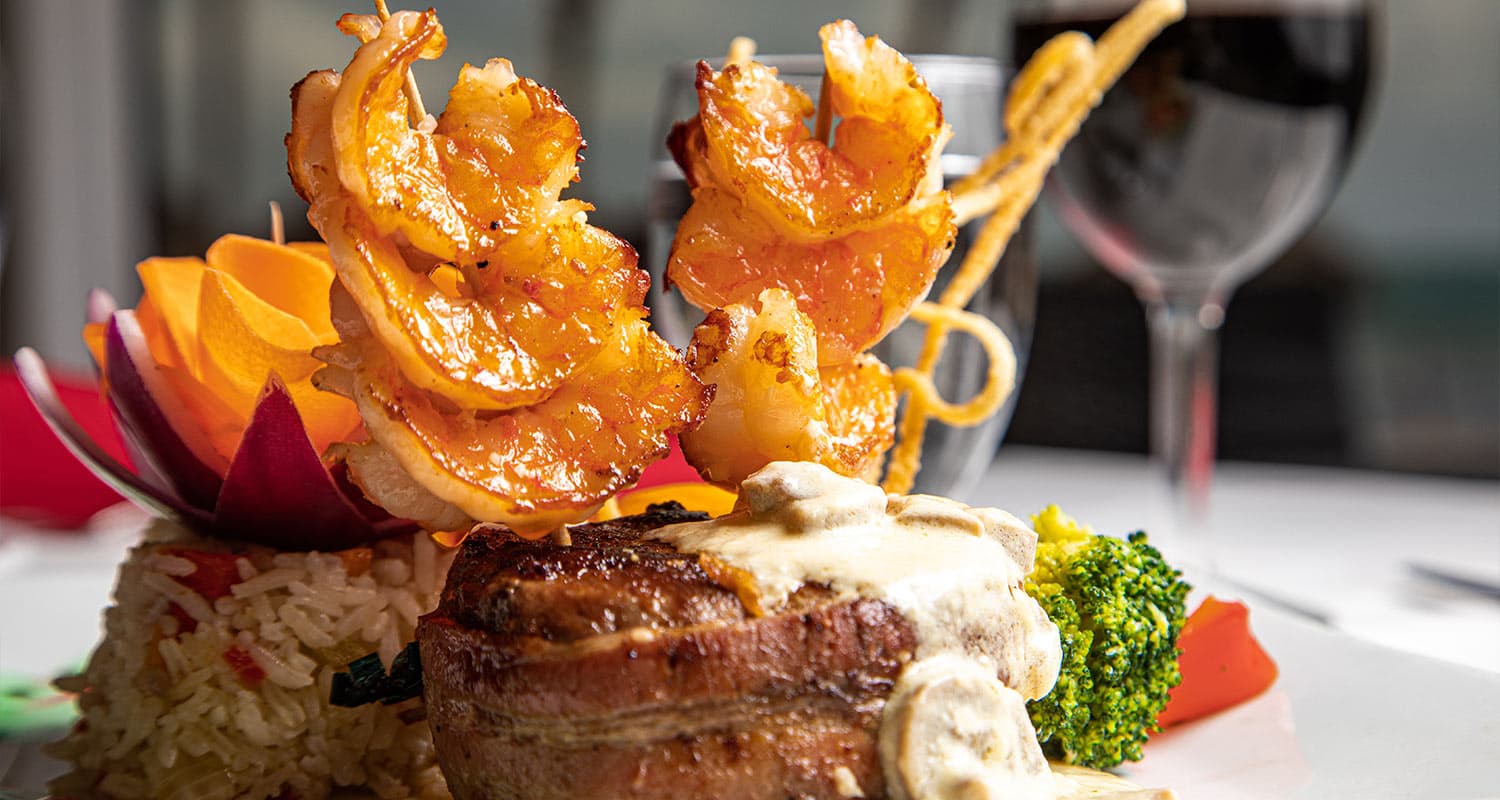 A gourmet dish that appears to be a steak and shrimp combo. The steak is wrapped in bacon and served with a creamy mushroom sauce. The shrimp are grilled and served on skewers.