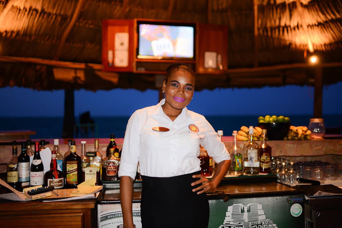 A tropical beach bar in the evening. A bartender in a white blouse smiles in front of the bar.