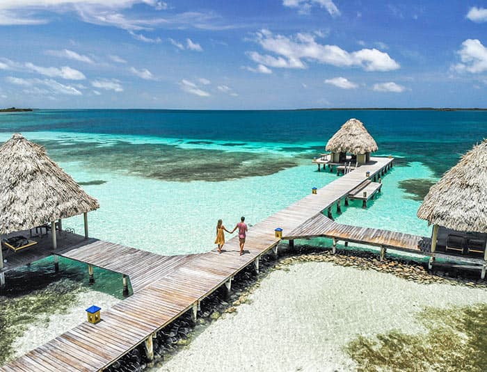 Couple walking on a wooden pier at Coco Plum Island Resort in Belize, with overwater bungalows and crystal-clear turquoise waters in the background.
