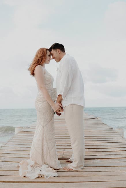A couple holds hands and leans towards each other on a wooden pier by the sea.