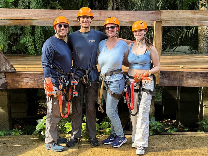 A group of four people wearing helmets and harnesses stands in front of a wooden structure in a lush, green environment.