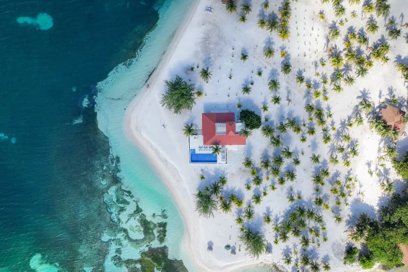 Aerial view of a tropical island featuring a house with a red roof surrounded by palm trees and clear blue waters.