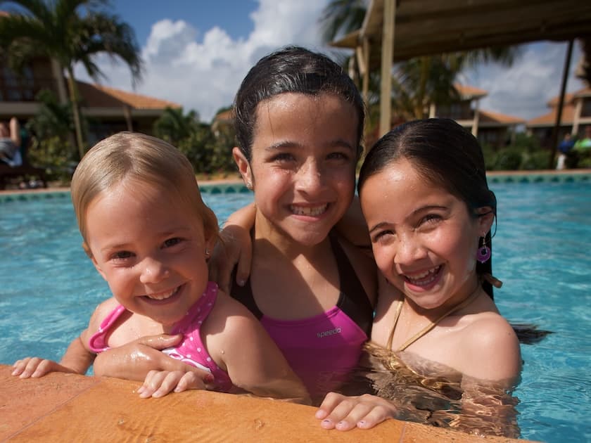 Three smiling girls play together in a swimming pool under a sunny sky.