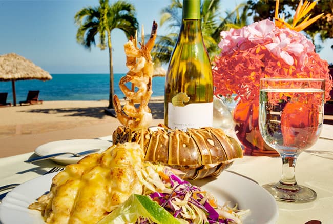 A gourmet seafood dish with lobster, shrimp, and vibrant slaw, accompanied by a bottle of wine and a glass, set against a beach backdrop.