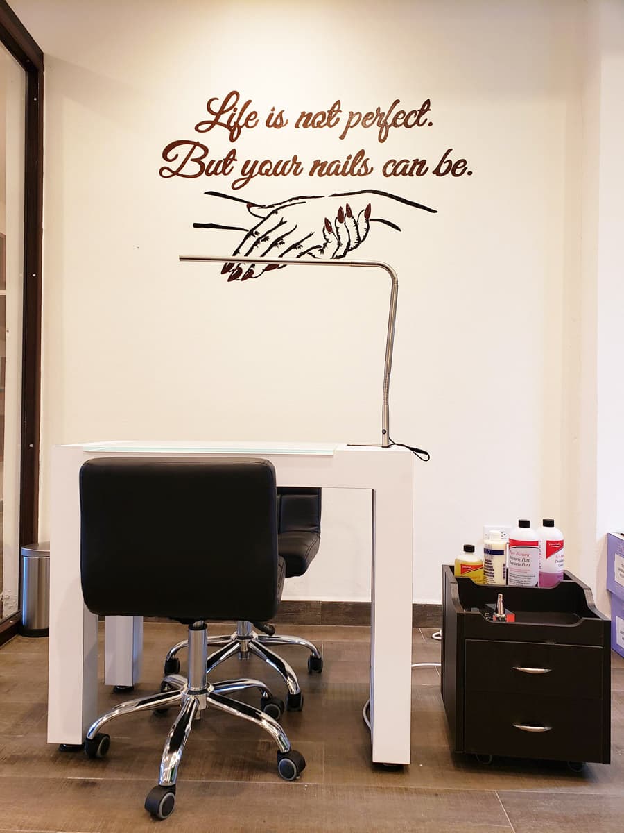 A nail salon station with a white table, two black swivel chairs, and a wall decal with the quote "Life is not perfect. But your nails can be." There is also a small cart with nail polish and other supplies. The overall atmosphere appears clean and modern.