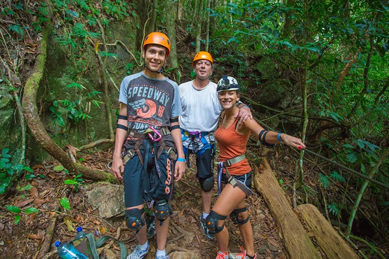 Three climbers in helmets and harnesses pose together in a lush, green forest. Three climbers in helmets and harnesses pose together in a lush, green forest.